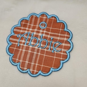 May include: A decorative patch with a scalloped edge and a plaid pattern in shades of orange and white. The name "Abbie" is embroidered in blue script. The patch has a light blue border.