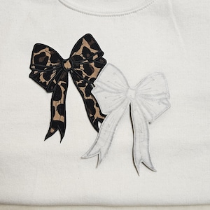 May include: A white sweatshirt with two iron-on patches. One patch is a leopard print bow and the other is a white bow.