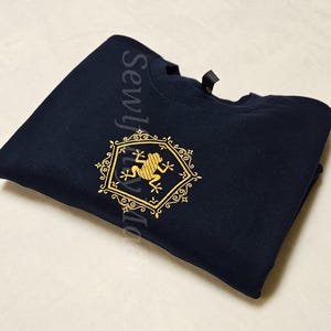 May include: A navy blue t-shirt with a gold embroidered design of a frog inside a hexagonal frame. The frame is decorated with intricate scrollwork.