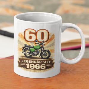 Könnte beinhalten: Weiße Keramik-Tasse mit einem Vintage-Motorrad-Motiv in Grün und dem Text "60 LEGENDÄR SEIT 1966" in Braun und Weiß. Das Design beinhaltet einen Retro-Sonnenstrahlen-Hintergrund.