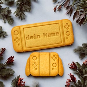 Op de afbeelding: Twee goudgele koekjes in de vorm van een gameconsole, één rechthoekig met de tekst "dein Name" en de andere een kleinere controller. De koekjes hebben knoopdetails en zijn omgeven door groen.