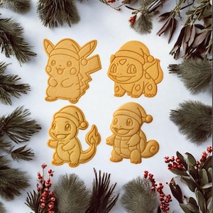 Könnte beinhalten: Vier hellbraune Kekse in Form von Pokémon-Charakteren, die jeweils einen Weihnachtsmannhut tragen. Zu den Charakteren gehören Pikachu, Bulbasaur, Charmander und Squirtle. Die Kekse sind auf einer weißen Oberfläche angeordnet, umgeben von Zweigen und roten Beeren.