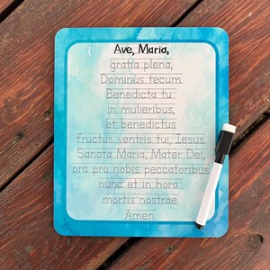 May include: A rectangular dry-erase board with a blue watercolor border. The board features the text of the Hail Mary prayer in a handwritten font. A white dry-erase marker with a black cap rests on the board.