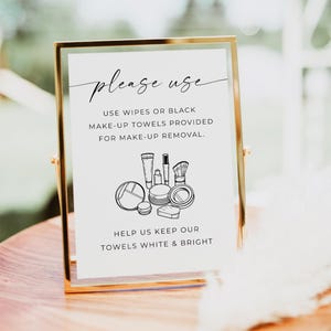 Op de afbeelding: Een goudkleurig bord met de tekst "please use" in elegant schrift. Het bord bevat instructies voor make-up verwijdering, met een afbeelding van make-up producten. De tekst moedigt het gebruik van doekjes of zwarte handdoeken aan.