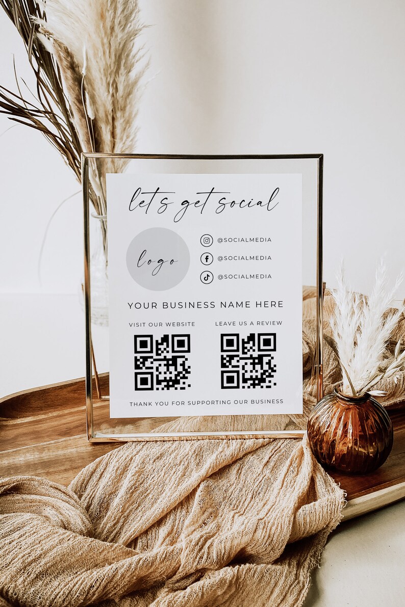 May include: A white sign with a gold frame, featuring the text "let's get social" and a logo placeholder. The sign also includes social media icons and QR codes for visiting the website and leaving a review. The text "YOUR BUSINESS NAME HERE" is included at the top of the sign. The sign is sitting on a wooden surface with a brown fabric and a vase with dried flowers.