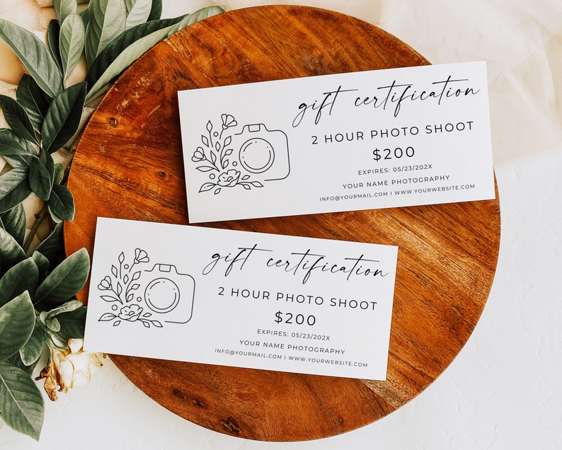 Op de afbeelding: Twee witte cadeaubonnen met zwarte tekst en een eenvoudige lijntekening van een camera met bloemen. De tekst luidt "gift certification 2 hour photo shoot &euro;200 EXPIRES: 05/23/202X YOUR NAME PHOTOGRAPHY INFO@YOURMAIL.COM WWW.YOURWEBSITE.COM"