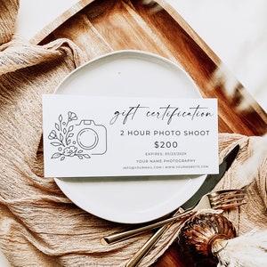 Op de afbeelding: Een wit cadeaubon met een zwarte lijntekening van een camera en bloemen. De tekst luidt "gift certification 2 hour photo shoot &euro;200 EXPIRES: 05/23/202X YOUR NAME PHOTOGRAPHY INFO@YOURMAIL.COM I WWW.YOURWEBSITE.COM"