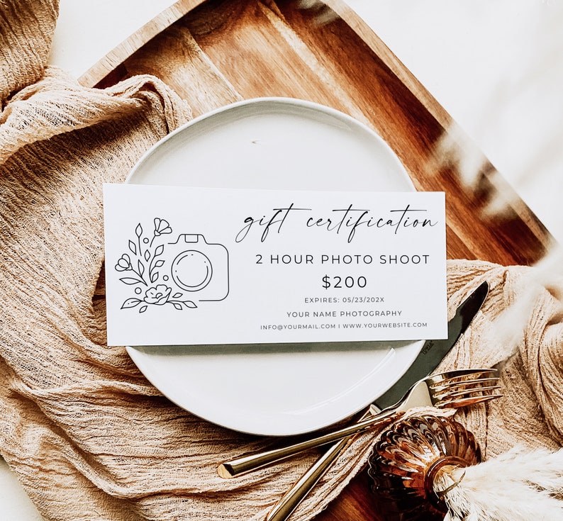 Op de afbeelding: Een wit cadeaubon met een zwarte lijntekening van een camera en bloemen. De tekst luidt "gift certification 2 hour photo shoot &euro;200 EXPIRES: 05/23/202X YOUR NAME PHOTOGRAPHY INFO@YOURMAIL.COM I WWW.YOURWEBSITE.COM"