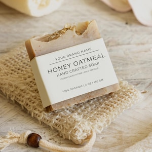 May include: A bar of Honey Oatmeal hand-crafted soap, with a white label that reads "Your Brand Name, Honey Oatmeal, Hand Crafted Soap, Vegan, Cruelty Free, Cold Pressed, 100% Organic, 113g | 150 gm." The soap rests on a beige, woven soap saver.