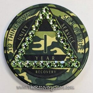 May include: A round, camouflage-patterned coin with a green triangle in the center, embellished with green rhinestones. The coin features the text "1 YEAR RECOVERY" and the phrases "TO THINE OWN UNITY" and "SELF BE TRUE SERVICE".