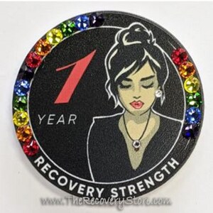 May include: A black circular medallion with a border of multi-coloured gemstones. The centre shows a woman's illustration, the number 1 in red, and the words "YEAR" and "RECOVERY STRENGTH".