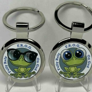 May include: Two round metal keychains with a cartoon frog design. One frog wears sunglasses, the other has large blue eyes. The keychains have the text "F.R.O.G. Fully Rely On God."
