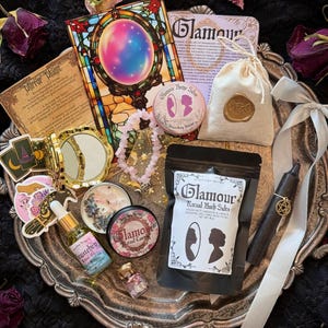 May include: A collection of mystical items on a silver tray, including a stained-glass mirror, a small bag with a gold seal, a black candle, and a bag of bath salts. Dried roses and a pink crystal necklace are also present.