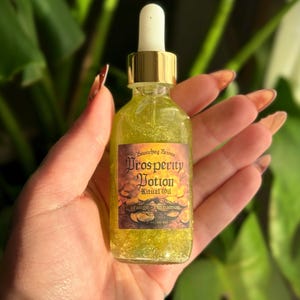 May include: A small glass bottle filled with a shimmering, golden liquid. The label reads "Prosperity Potion Ritual Oil" with "orange-cinnamon" below. The bottle has a gold cap and dropper, and is held in a hand.