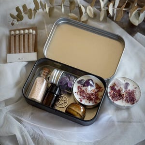 May include: A small metal tin filled with various items for a ritual or spell. The tin contains a small bottle of sand, a bottle of essential oil, a small jar of amethyst crystals, two tealight candles with dried flowers and crystals, a small pentacle charm, and a tiger's eye stone.