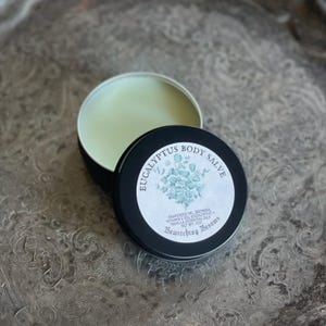 May include: A black tin with a white label that reads "Eucalyptus Body Salve" with a drawing of eucalyptus leaves. The label also lists the ingredients: grapeseed oil, beeswax, vitamin E oil, eucalyptus and vanilla essential oils. The tin is 2 oz. and the brand is Bewitching Besoms.