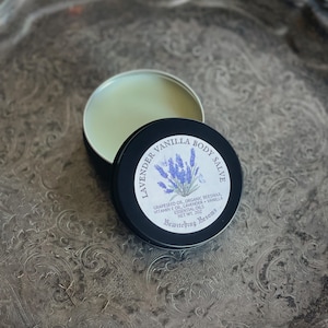May include: A black tin container with a white label that reads "Lavender Vanilla Body Salve". The label also includes a small illustration of lavender flowers and the text "Grapeseed Oil, Organic Beeswax, Vitamin E Oil, Lavender + Vanilla Essential Oils, Net Wt. 2 oz, Bewitching Besoms".