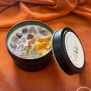 May include: A black tin candle with a white label that reads "Lemon & Lavender Soy Candle Ali." The candle is topped with a white wax layer and decorated with purple crystals and dried pineapple pieces.