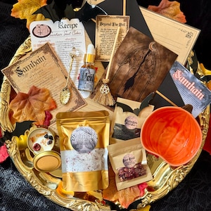 May include: A Samhain-themed ritual kit on a gold tray featuring a pumpkin-shaped bowl, a small dropper bottle labeled Beyond the Veil, a gold spoon, a miniature broom, pouches of black salt and rose petals, a gold pouch of bath salts, and cards.