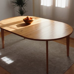Könnte beinhalten: Ein ovaler Esstisch aus hellem Holz mit natürlicher Oberfläche. Der Tisch hat eine Einlegeplatte und steht auf vier schlanken Beinen. Der Tisch steht auf einem hellbeigen Teppich.