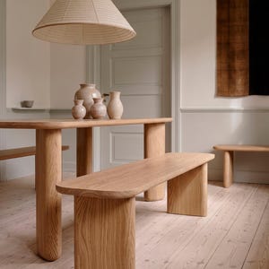 May include: A light wood dining table and benches set in a room with a light-colored wood floor. A large, beige, cone-shaped pendant light hangs above the table. Decorative vases sit on the table.