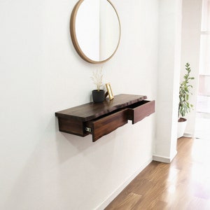 Può includere: Un ripiano a muro in legno marrone scuro con due cassetti, uno aperto e uno chiuso. Il ripiano è montato su una parete bianca e ha uno specchio rotondo dorato sopra.