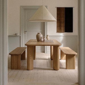 Könnte beinhalten: Ein Esstisch aus hellem Holz mit abgerundeten Beinen und zwei passenden Bänken. Über dem Tisch hängt eine große, konische Pendelleuchte. Dekorative Vasen und ein kleiner Stapel Bücher stehen auf dem Tisch. Der Raum hat hellfarbene Wände und einen Holzboden.