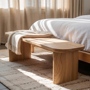 Può includere: Panca in legno chiaro con piano arrotondato e gambe rettangolari. Una coperta color crema con frange è drappeggiata sulla panca. La panca è in una camera da letto, vicino a un letto e un tappeto.