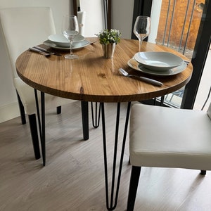 Vista de la mesa redonda con patas hairpin. Hay dos sillas blancas colocadas y también dos platos y dos copas para vino. También hay cubiertos para comer y una maceta pequeña.