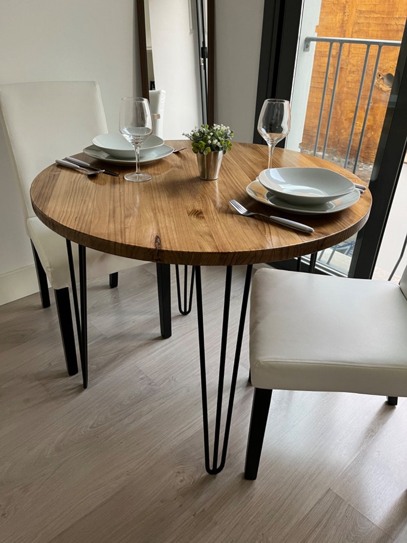 Vista de la mesa redonda con patas hairpin. Hay dos sillas blancas colocadas y también dos platos y dos copas para vino. También hay cubiertos para comer y una maceta pequeña.