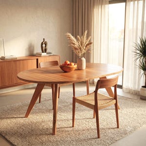 Peut inclure: Une table à manger ovale en bois avec des pieds inclinés et deux chaises assorties dotées d'assises tressées. La table porte un bol en bois rempli d'oranges et un vase contenant des herbes de la pampa séchées, dans un intérieur clair.