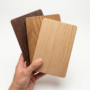 Op de afbeelding: Een hand houdt drie rechthoekige houten stalen in verschillende bruin- en beigetinten vast. De houtnerf is zichtbaar, met kleuren variërend van donkerbruin tot licht eiken. De stalen hebben afgeronde hoeken.