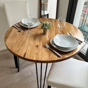 Mesa redonda de madera hecha a mano, con patas hairpin y para 2 personas.
El color de la madera es castaño.
Perspectiva desde arriba.
La luz entra por una ventana a la derecha.
En la mesa hay dos platos y dos copas. También un maceta en el centro.