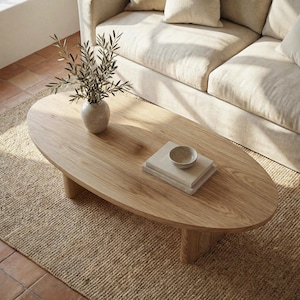 Peut inclure: Table basse ovale en bois clair, avec un vase contenant de la verdure, des livres et un petit bol. La table est posée sur un tapis tissé, avec un canapé beige en arrière-plan. La table mesure environ 127 cm de long.