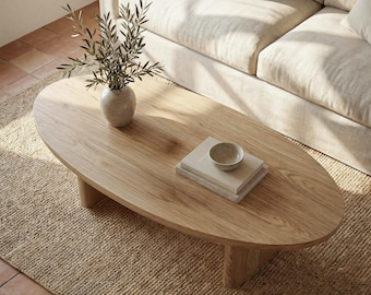 May include: Oval wooden coffee table with a light finish, featuring a vase with greenery, books, and a small bowl. The table is set on a woven rug, with a beige sofa in the background. The table is approximately 50 inches long.