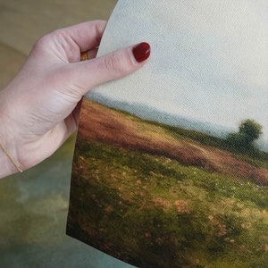 May include: A close-up of a canvas print depicting a landscape scene. The print shows a grassy field with a distant horizon and a small tree in the foreground. The sky is a light blue with some clouds.