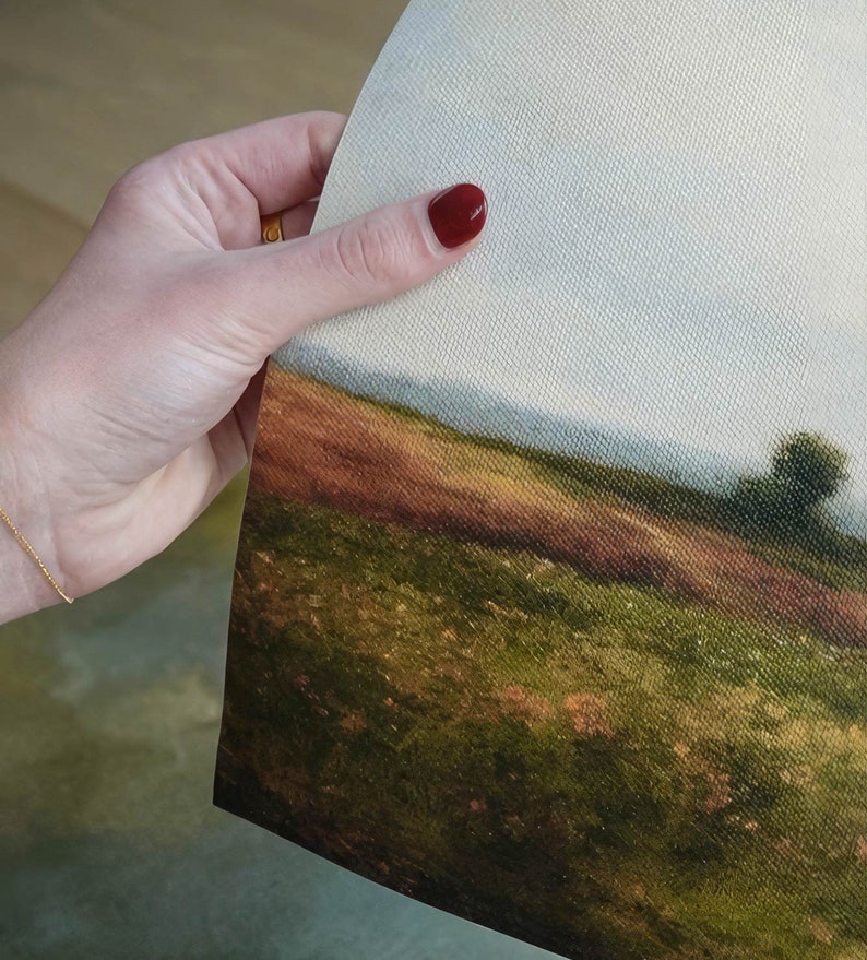 May include: A close-up of a canvas print depicting a landscape scene. The print shows a grassy field with a distant horizon and a small tree in the foreground. The sky is a light blue with some clouds.