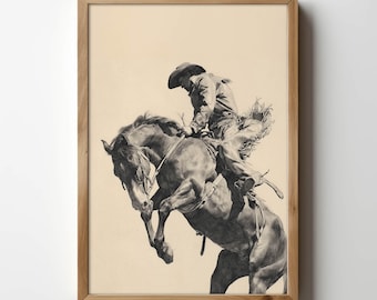 May include: A framed black and white print of a cowboy riding a bucking bronco. The cowboy is wearing a hat and chaps, and the horse is rearing up. The print is in a light brown wooden frame.