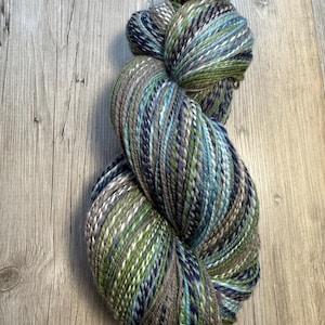 May include: A skein of variegated yarn in shades of green, blue, gray, and white. The yarn is tightly wound and appears soft, with a textured surface. The color palette evokes a natural, earthy feel, suitable for knitting or crochet projects.