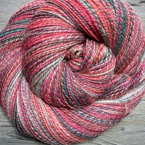 May include: A large skein of colorful yarn, predominantly pink and red, with accents of gray, white, and teal. The yarn is tightly wound, showcasing a textured appearance. The image is taken on a wooden surface.