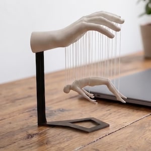 May include: A sculpture featuring a matte white hand mounted on a black metal stand, with a small white human figure suspended beneath the fingers by thin white strings, creating a marionette-like effect in a monochromatic design.