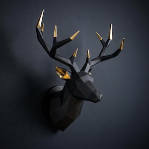 Könnte beinhalten: Eine geometrische Wandskulptur in Form eines Hirschkopfes mit matter schwarzer Oberfläche und kontrastierenden metallisch goldenen Akzenten an den Geweihspitzen und den Innenohren, bestehend aus scharfen, kantigen Facetten.