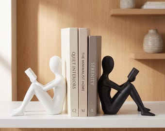 May include: A pair of bookends featuring two abstract human figures, one white and one black, each sitting and holding a small book. Between the figures are three books with spines labeled Quiet Interiors, The Minimalist Home, and Serenity.