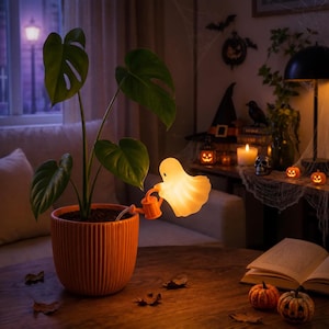 Op de afbeelding: Een monstera-plant in een geribbelde oranje keramische pot met een kleine, oplichtende, witte spookvormige lamp bevestigd aan een miniatuur oranje gieter. De spooklamp verspreidt een warm, zacht licht over de groene bladeren van de plant.