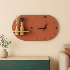 May include: An oval-shaped terracotta wall clock featuring black hands and numerals at 3, 6, 9, and 12. A small attached wooden shelf holds two beige vases, one containing dried pampas grass and the other holding a small bunch of purple flowers.