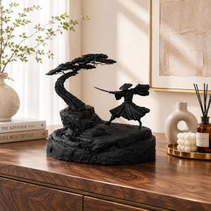 May include: A matte black sculpture featuring a warrior figure in a wide-brimmed hat holding a sword, positioned on a rocky base next to a stylised tree. The entire piece is monochromatic black with detailed textures on the rock and tree.