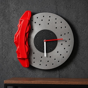 Puede incluir: Un reloj de pared diseñado para parecerse a un disco de freno plateado circular con una pinza de freno roja unida al lado izquierdo. El centro presenta una placa circular negra con una manecilla de hora roja, una manecilla de minutos blanca y un segundero rojo.