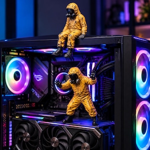 May include: Two yellow hazmat suit figurines with black gas masks and boots positioned inside a black computer case. The case features internal components illuminated by vibrant, multi-colored RGB lighting, including circular fans and a graphics card.