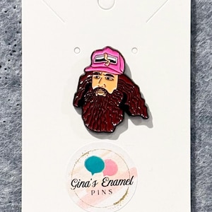 May include: A pink and black enamel pin featuring a cartoon illustration of a man with a long brown beard wearing a pink baseball cap with a black logo. The pin is on a white card with the text "Gina's Enamel Pins".