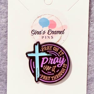 May include: A pin with a cross design in light blue and black, with the words "Pray on it, Pray over it, Pray through it." The pin is on a white card with the text "Gina's Enamel Pins."